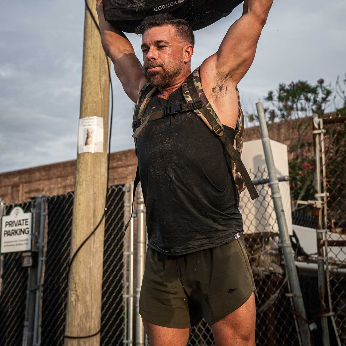 A man wearing a black tank top and GORUCK's Men’s Ranger Panties - ToughStretch in green is lifting a heavy sandbag overhead. He stands outdoors near a fenced area with an industrial backdrop, while the cloudy sky looms above. A Private Parking sign nearby evokes the assured resilience of the ToughStretch fabric.