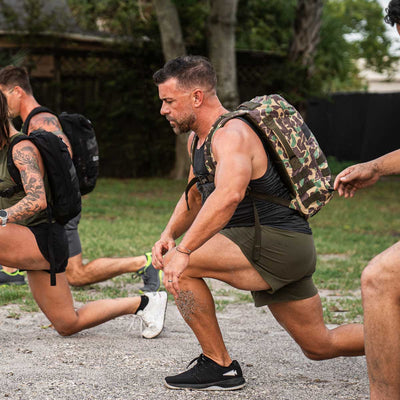 In a grassy tree-lined area, several people are exercising outdoors, each doing lunges with backpacks. The focus is on a man in the foreground wearing a black tank top and camo backpack, sporting GORUCK’s Men’s Ranger Panties made from ToughStretch fabric, providing both flexibility and comfort.