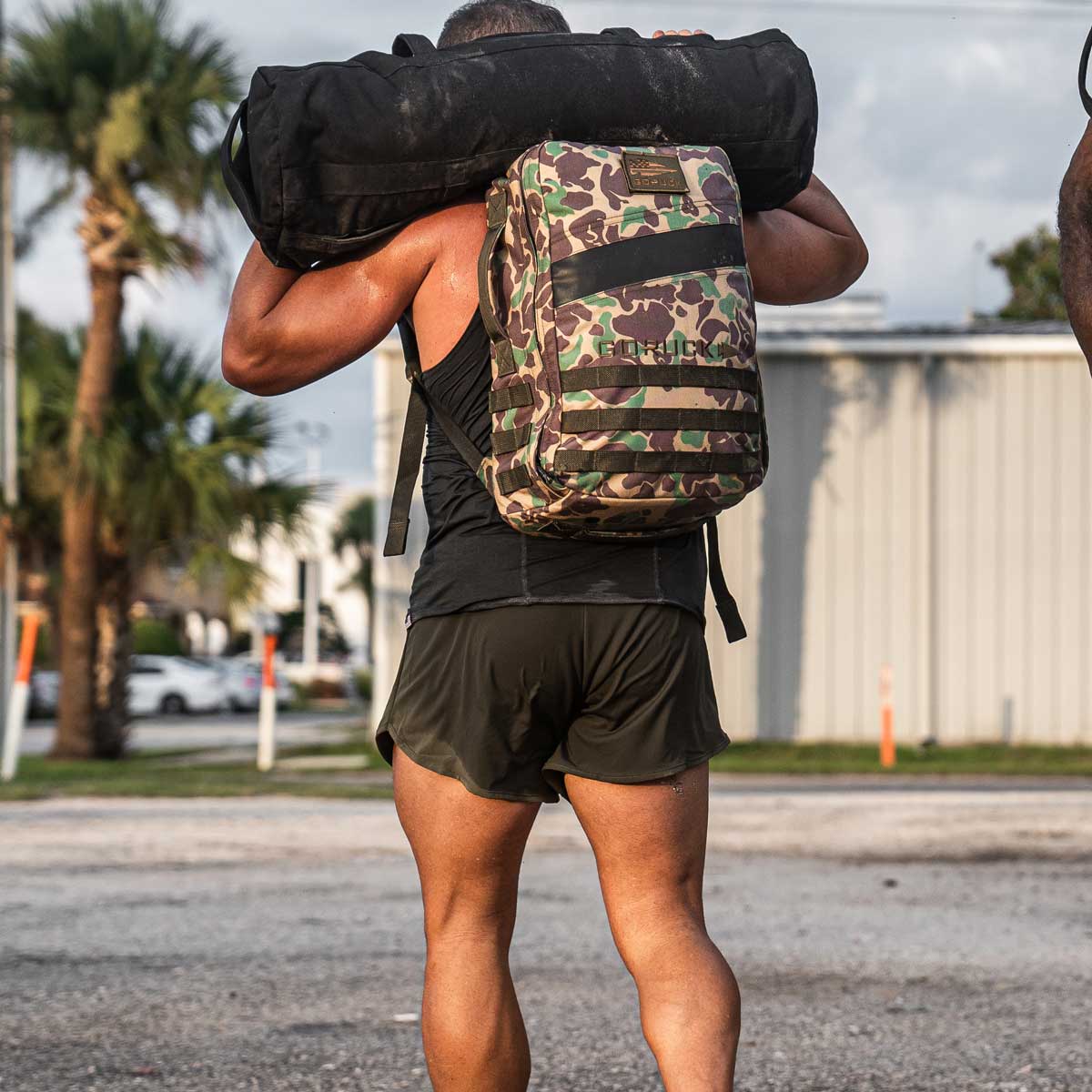 A person wearing GORUCK's Men’s Ranger Panties, made with ToughStretch fabric, carries a camo-pattern backpack and a large black duffel bag outdoors. The scene is set against a backdrop of palm trees and buildings, symbolizing adventures supported by the Scars Lifetime Guarantee.