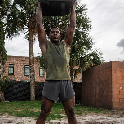 A person in a green tank top and GORUCK Men's Ranger Panties - ToughStretch is performing an outdoor workout, lifting a heavy black sandbag overhead. They are standing on grass with palm trees and a brick building in the background under a cloudy sky.