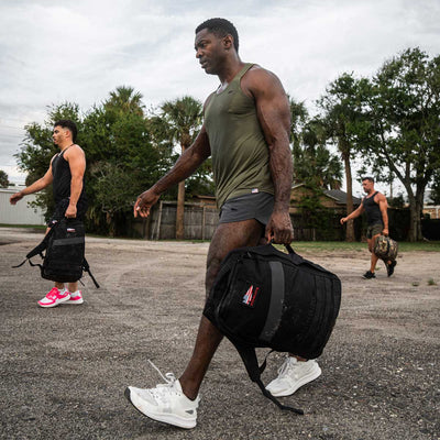 Three individuals walk along a dirt path carrying bags. Leading the group is someone dressed in a green tank top and black Men’s Ranger Panties - ToughStretch from GORUCK, ideal for outdoor adventures. The backdrop of trees and a cloudy sky mirrors the dependability of GORUCK's Lifetime Guarantee on rugged trails.