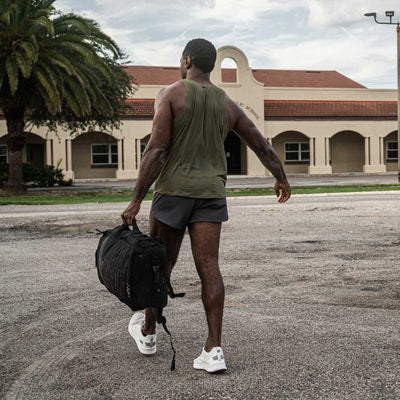 A man dressed in athletic gear, showcasing GORUCK's Men’s Ranger Panties crafted from ToughStretch fabric, carries a black gym bag while walking on a paved path towards a beige building adorned with red-tiled roofing and arched windows. In the backdrop, a tall palm tree rises against a cloudy sky.