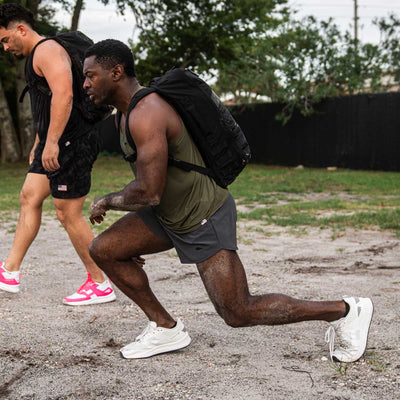 Two men in athletic gear, each with a backpack, perform lunges on a sandy outdoor surface. They appear focused, one man wearing a green tank top and gray GORUCK Men’s Ranger Panties featuring ToughStretch fabric, while the other is dressed in black. Trees and a fence are visible in the background.