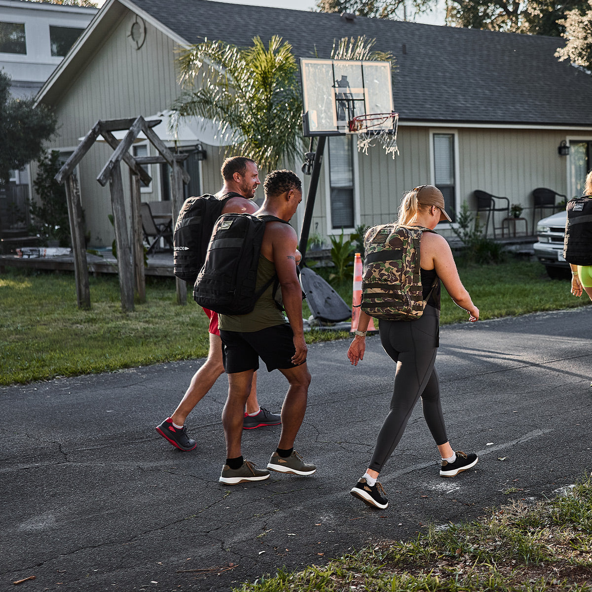 A group of four people strolls along a suburban street, each carrying backpacks made of CORDURA Ballistic Nylon. They pass a wooden swing set and basketball hoop in front of a light-colored house with a sloping roof, while their GORUCK Men's Ballistic Trainers - Ranger Green + White W / Coyote Reflective Spearhead navigate the sunlit pavement with enhanced support.