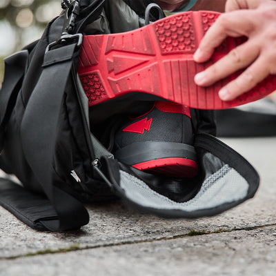 A person is adding or taking out the Men's Ballistic Trainers from a black duffel bag. These GORUCK sneakers, in Wolf Grey with red reflective details, are made from CORDURA® Ballistic Nylon and rest open on a concrete surface outdoors.