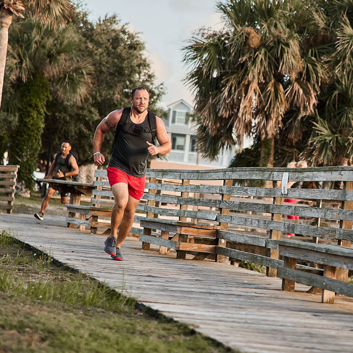 A man in a black tank top and red shorts runs on a wooden boardwalk lined with palm trees, sporting Men's Ballistic Trainers in Wolf Grey with High Risk Red and a Red Reflective Spearhead by GORUCK. Another person follows behind, also wearing functional fitness footwear. A house stands in the background under a cloudy sky.