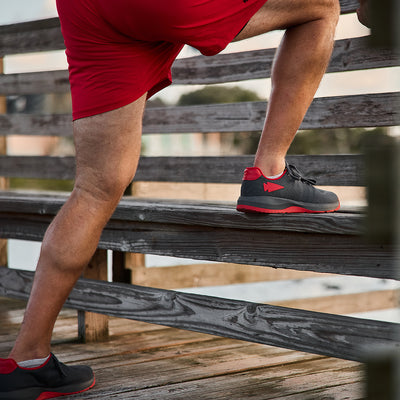 A person wearing GORUCK's Men's Ballistic Trainers in Wolf Grey and High Risk Red with a Red Reflective Spearhead, along with red shorts, is stretching one leg on a wooden bench outdoors against a blurred scenic background.