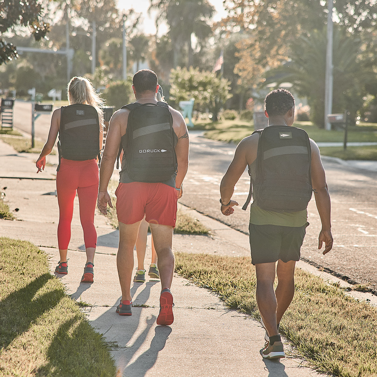 Three individuals are walking along the sidewalk, all wearing GORUCK Men's Ballistic Trainers in Wolf Grey with High Risk Red featuring a Red Reflective Spearhead and carrying backpacks. They are dressed in athletic gear, with the two on the left sporting red outfits and the person on the right clad in green and black. Behind them, trees line a nearby road.