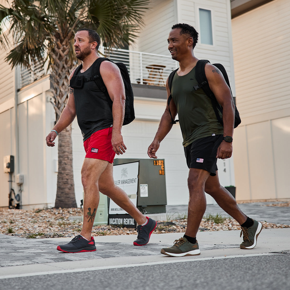 Two men are walking briskly on a sidewalk in a residential area, clad in athletic clothing and backpacks crafted from CORDURA® Ballistic Nylon. They sport GORUCK Men's Ballistic Trainers in Wolf Grey and High Risk Red with Red Reflective Spearhead, perfect for functional fitness footwear. One has a tattoo on his leg, while palm trees and houses line the background.