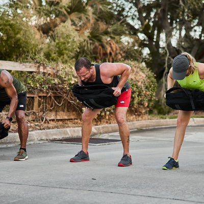 Three individuals engage in outdoor exercise, bending over with weighted bags in hand. They are outfitted in athletic wear and GORUCK's Men's Ballistic Trainers in Wolf Grey with High Risk Red accents and a Red Reflective Spearhead. They stand on a paved surface surrounded by lush greenery.