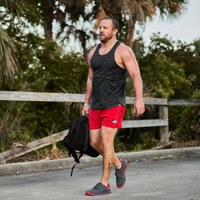 Wearing a black tank top and red shorts, the man walks along a path carrying a backpack, highlighting his Men's Ballistic Trainers in Wolf Grey and High Risk Red with Red Reflective Spearhead by GORUCK. His muscular build and tattoos on his left leg stand out against the lush greenery and wooden fence backdrop.