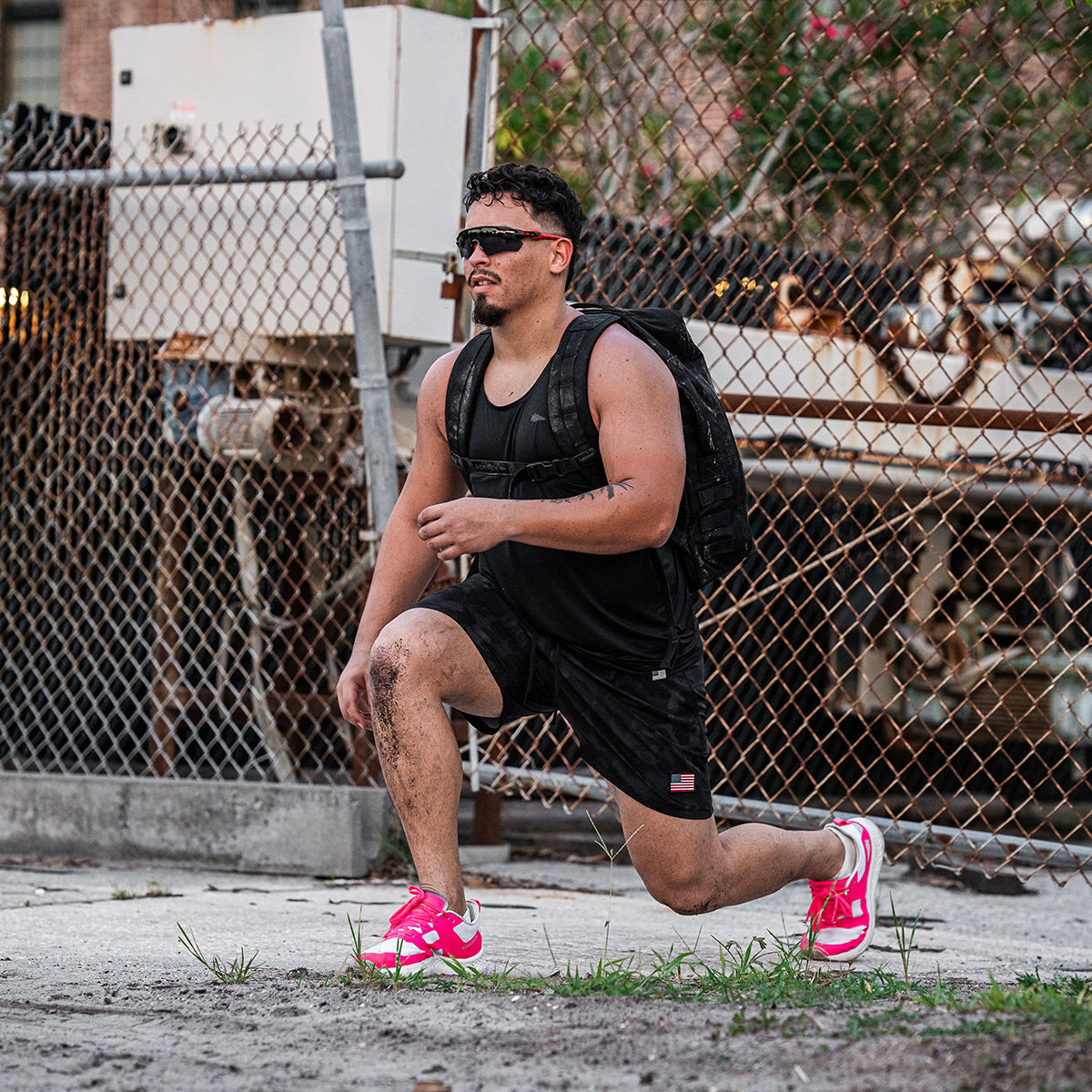 Dressed in a black outfit and wearing the Men's Rough Runner - Hot Pink by GORUCK, this individual performs a lunge outdoors. They carry a backpack and are surrounded by industrial equipment and a chain-link fence, capturing the essence of a Rough Runner challenge on concrete terrain.