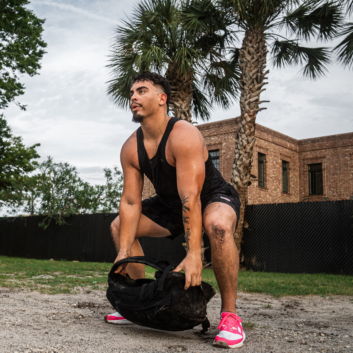 Wearing the Men's Rough Runner in Hot Pink by GORUCK, a person in a black tank top performs a squat lift with a sandbag outdoors. Palm trees and a brick building set the scene, perfectly capturing the spirit of Rough Runner.