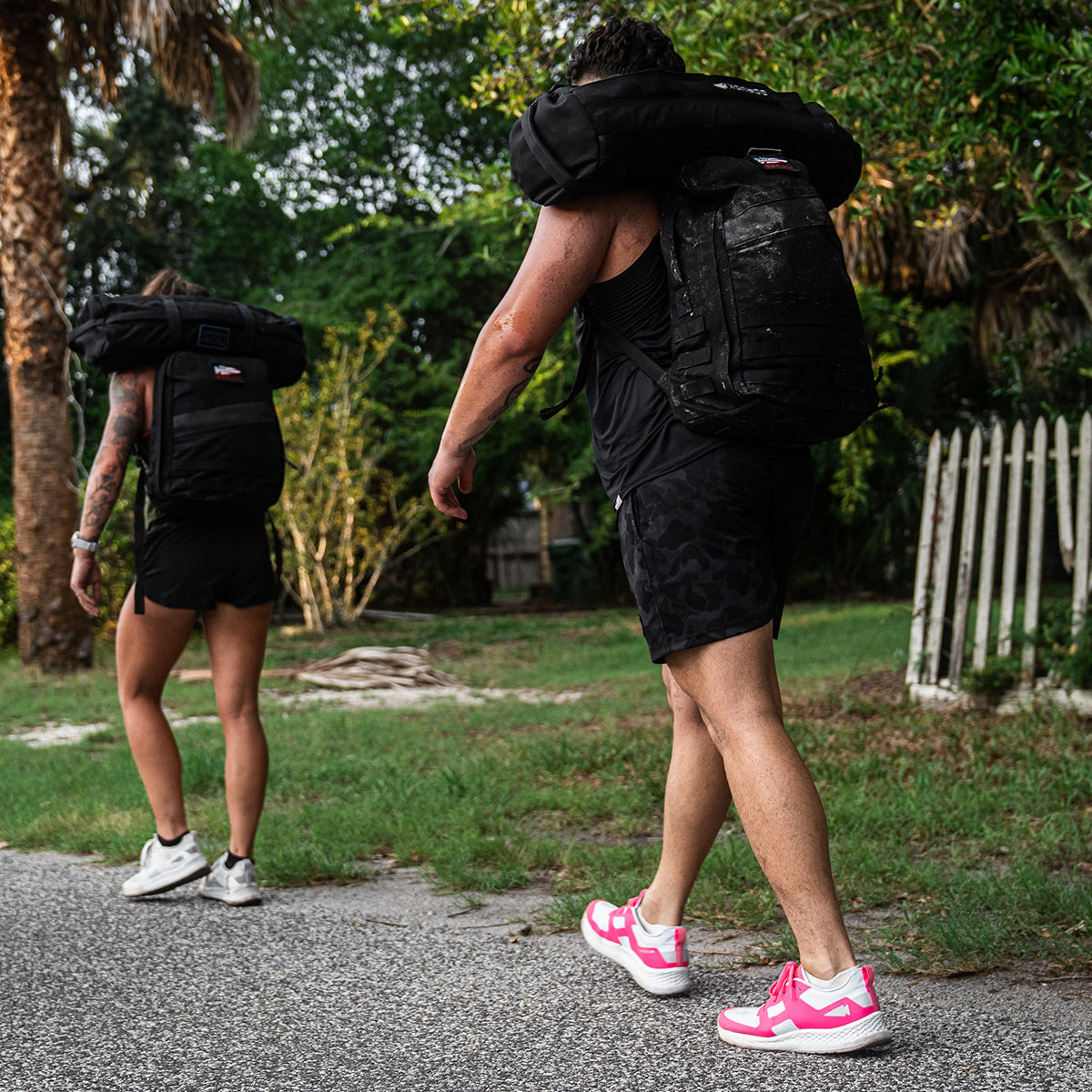 Two people stroll along a sidewalk, each with a Men's Rough Runner - Hot Pink sandbag by GORUCK slung over their shoulders. They wear sporty attire and running shoes, prepared for any challenge ahead. In the background, trees and a white picket fence evoke the feel of a Rough Runner course.