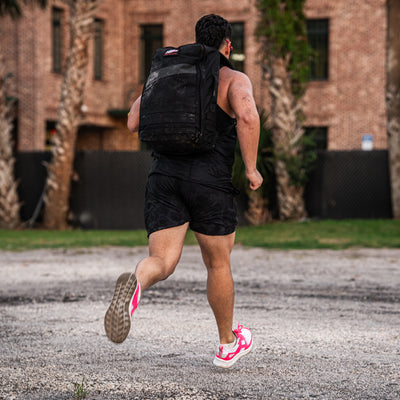 Sporting sleek Men's Rough Runner shoes in hot pink by GORUCK, an athlete in full gear strides along the gravel path. A large backpack bounces rhythmically on their back as they navigate past a brick building, flanked by palm trees and a sturdy fence in the background.