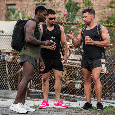 Three men in GORUCK Men's Rough Runner gear, likely getting ready for a Rough Runner event, stand outside by a chain-link fence, each holding an energy drink. One gestures animatedly while the others listen with focus. They're equipped with backpacks and running shoes, surrounded by lush greenery in the background.