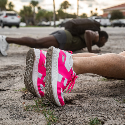 Close-up of a person doing push-ups on sandy ground, sporting vibrant pink and white Men's Rough Runner shoes in Hot Pink from GORUCK. Another person is in the background, mirroring the exercise. Parked cars and palm trees blur into the gradient density of the distant backdrop.