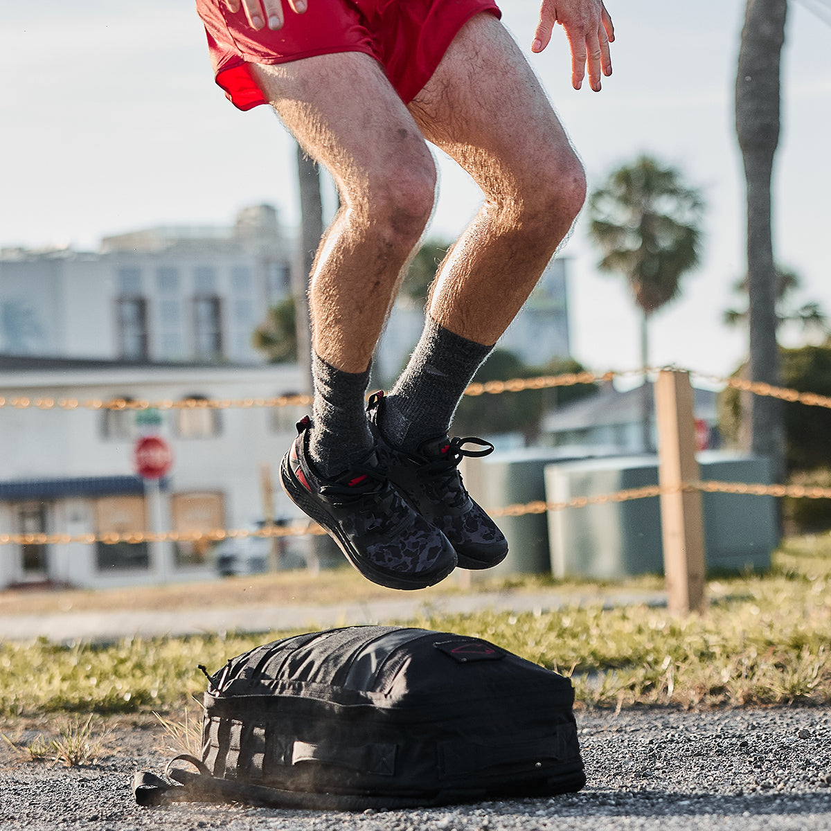 A person sporting the Men's Rough Runner in Midnight Frogskin and High Risk Red from GORUCK effortlessly leaps over a black backpack on a gravel path. These high mileage running shoes grip the ground with ease. In the background, palm trees and buildings blur against a clear sky, crafting an idyllic beach setting ideal for any Rough Runner adventure.