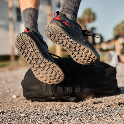 A Men's Rough Runner - Midnight Frogskin + High Risk Red by GORUCK lands on a black duffle bag, their gray socks nestled in high mileage running shoes featuring a Gradient Density EVA Midsole. Dust swirls around the gravel ground, with blurred palms and buildings in the background, as red accents flash against the black footwear canvas.