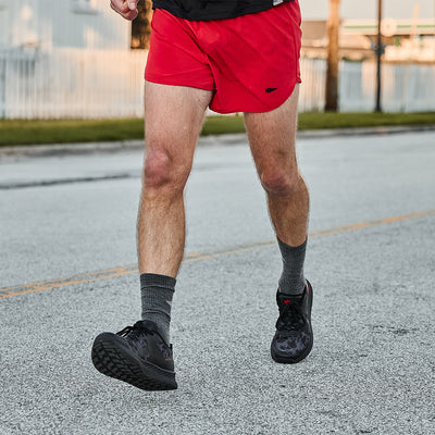 A GORUCK Men's Rough Runner - Midnight Frogskin + High Risk Red jogs down the street, sporting red shorts, dark socks, and sleek black running shoes with a Gradient Density EVA Midsole. In the background, a white fence lines the path alongside lush trees, perfectly complementing their high-mileage design for endurance.