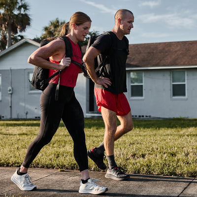 A woman and a man are walking together on a suburban sidewalk, wearing athletic clothing and carrying backpacks. The man steps lightly in his GORUCK Men's Rough Runner shoes in Midnight Frogskin + High Risk Red on the pavement. The sky is clear, with palm trees swaying near a house in the background.