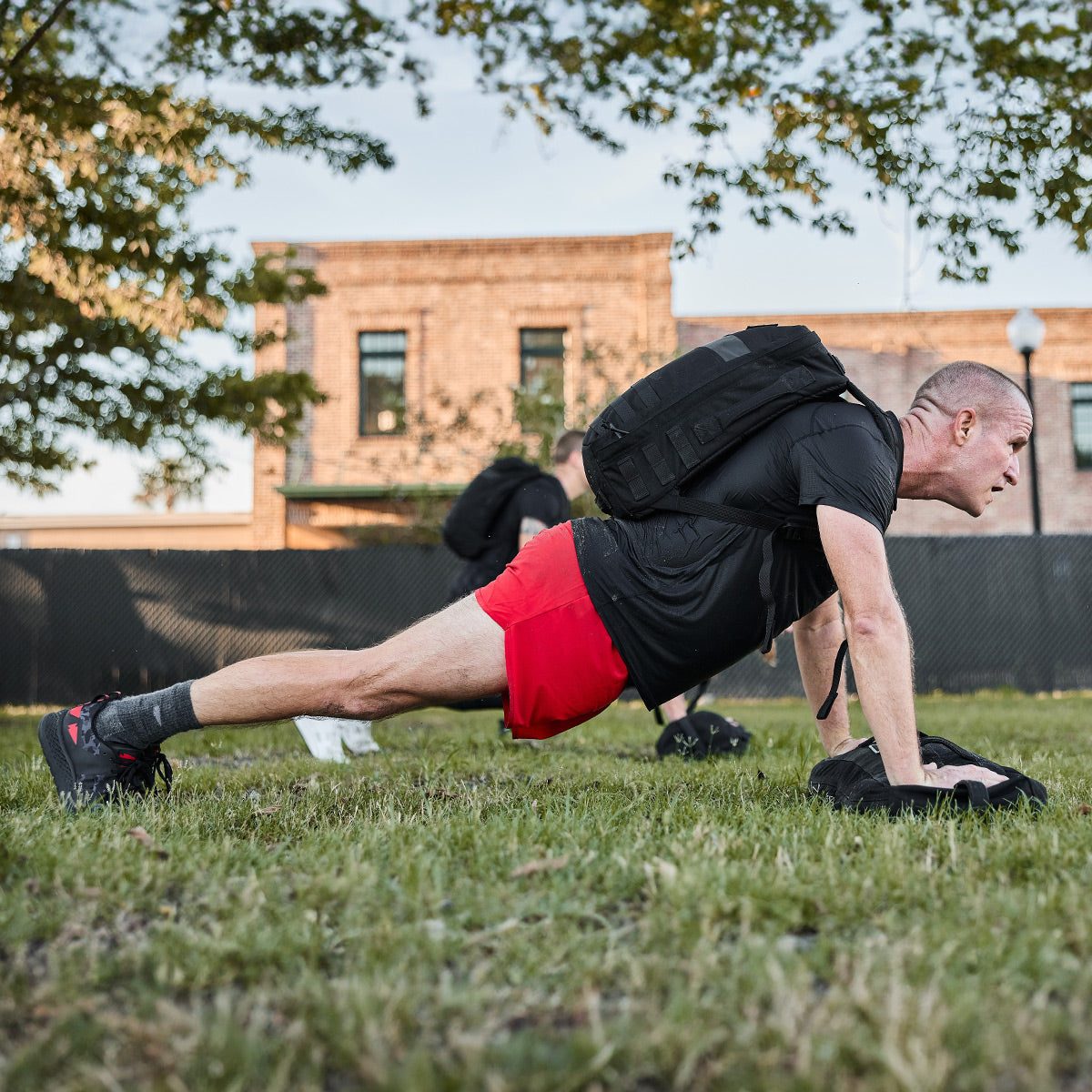 A person wearing GORUCK's Men's Rough Runner in Midnight Frogskin and High Risk Red performs a plank exercise on a grassy field. Dressed in a black shirt, red shorts, and gray socks, they maintain a steady form while buildings and trees are visible against the backdrop of a clear sky.