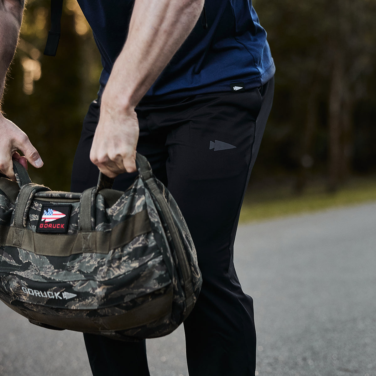 A person wearing GORUCK's Men’s Performance Joggers, designed with performance fabric for ultimate comfort, lifts a camouflage-patterned GORUCK bag on a road surrounded by greenery.