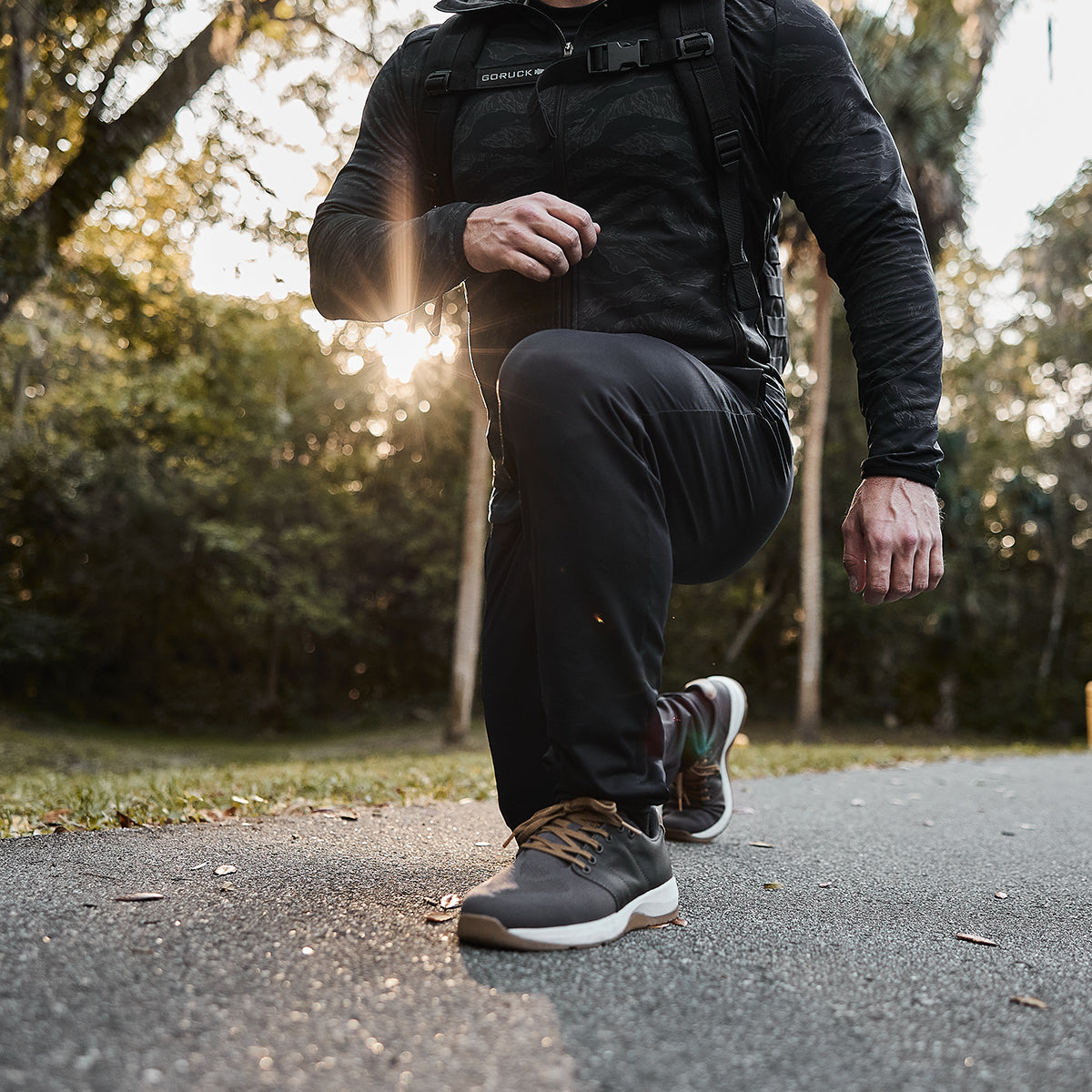 A person wearing Men’s Performance Joggers by GORUCK kneels on a paved path surrounded by lush green trees, with sunlight streaming through. The focus is on their shoes and the natural setting, evoking a moment of stretching or exercise in breathable performance fabric.