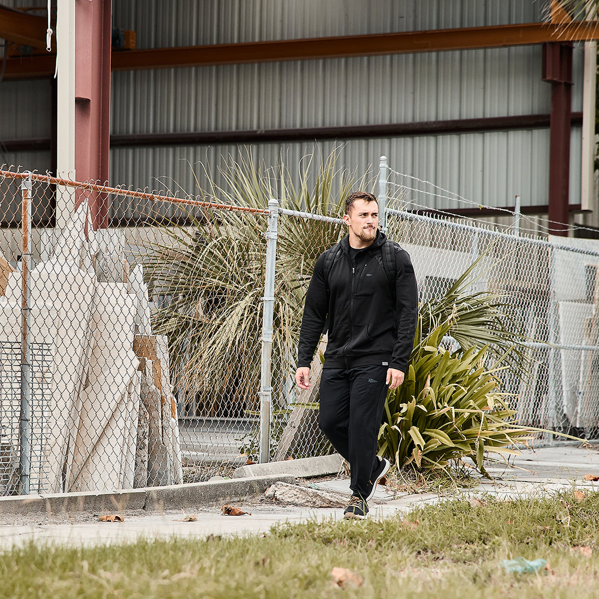 A man wearing GORUCK Men’s Performance Joggers strolls past a chain-link fence next to a metal building. He's carrying a backpack and walks by some grassy plants, with construction materials partially visible in the background.