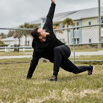 Dressed in men’s performance joggers by GORUCK, a person executes a dynamic stretch on a grassy field, extending their left arm upwards while kneeling on the ground. A chain-link fence and several buildings can be seen in the background.