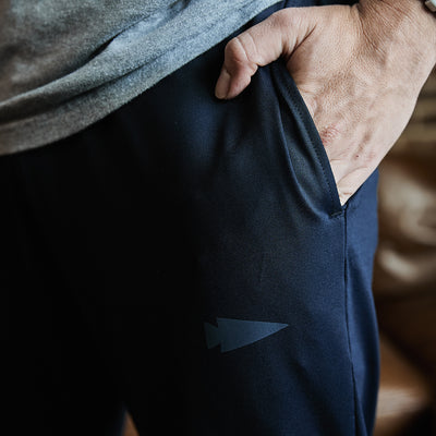 A person wearing GORUCK's Men's Performance Joggers, featuring a small arrowhead logo on the thigh, casually rests their hand in the pocket. They are also dressed in a light gray top crafted from moisture-wicking performance fabric.