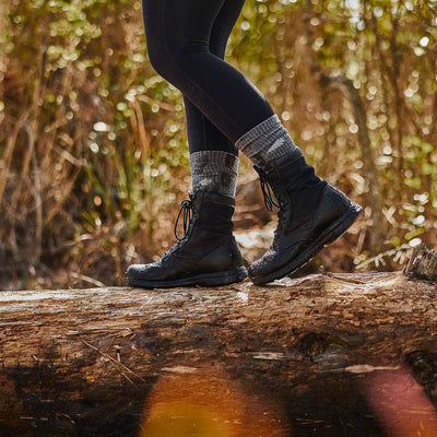 In a serene forest setting, a person stands gracefully on a log, wearing black hiking boots paired with GORUCK's Merino Challenge Socks - Crew in grey. Sunlight filters through the trees, creating an atmosphere as enduring as Scars' Lifetime Guarantee.