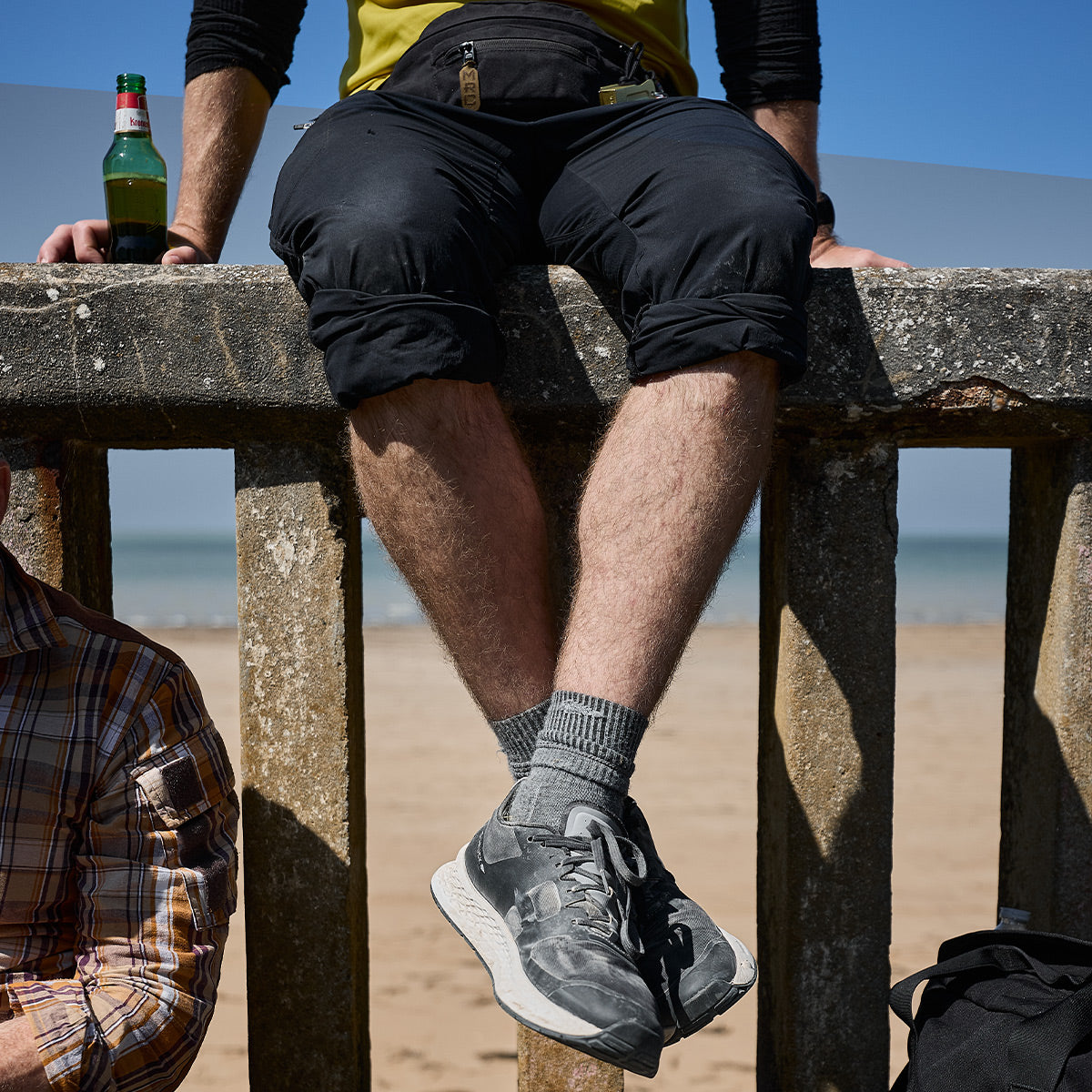 A person in rolled-up black pants sits on a weathered concrete wall at the beach, their legs dangling. They sport GORUCK Merino Challenge Socks - Crew in gray and sneakers. A closed green bottle and a fanny pack rest on the ledge beside them. Behind them, sand and sea bask under a blue sky.