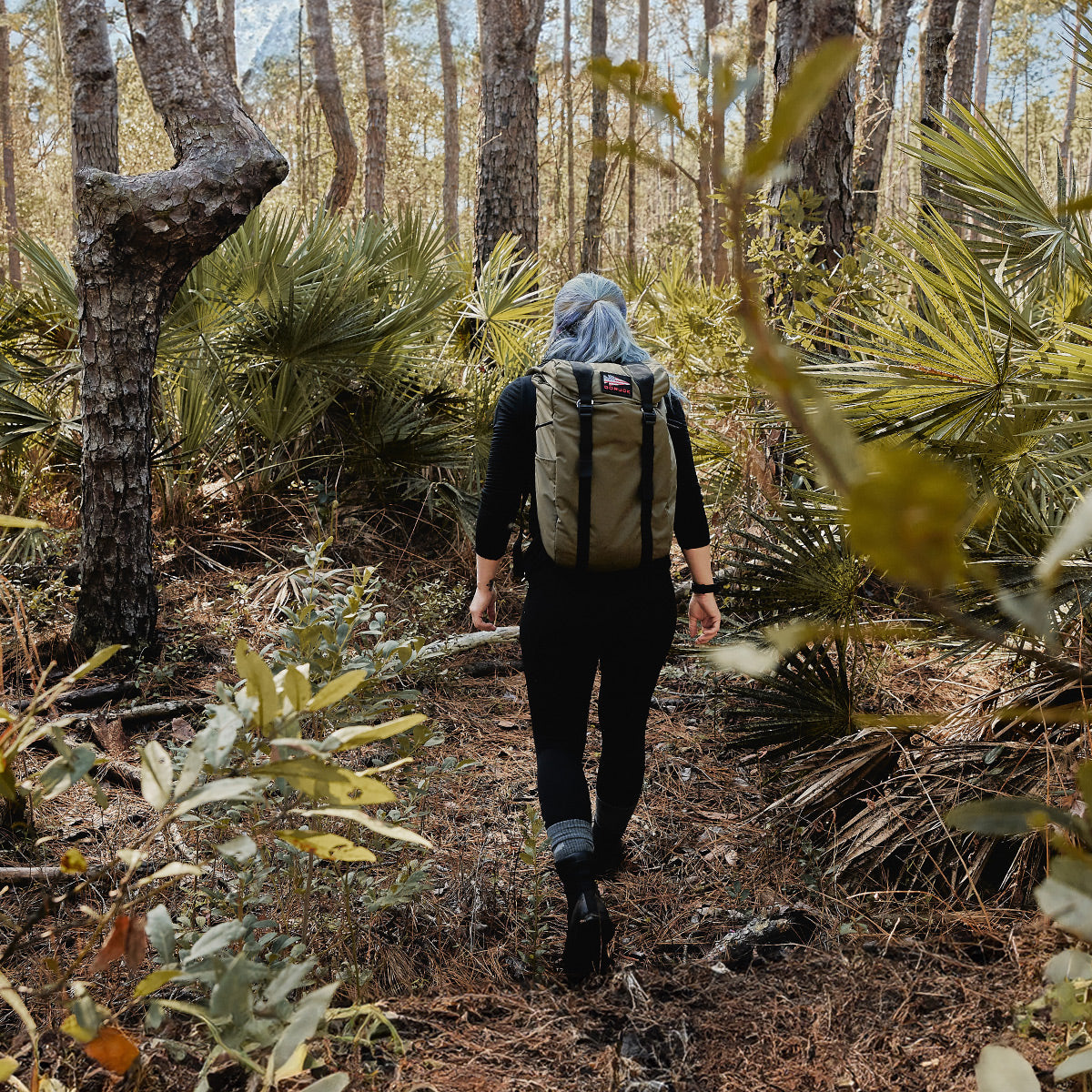 A person with a backpack walks through a dense forest, surrounded by tall trees and lush green foliage. Clad in GORUCK Merino Challenge Socks - Crew that promise comfort, they trek along the leaf-covered ground as sunlight filters through the branches above.