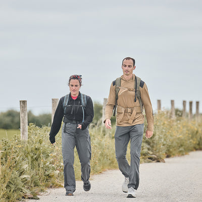 Two people enjoy a hike on a pathway surrounded by greenery. The person on the left wears a black top and gray pants, while the one on the right sports a tan jacket and gray pants. Both have backpacks, possibly carrying essentials like GORUCK's Merino Challenge Socks - Crew. They trek under a cloudy sky.