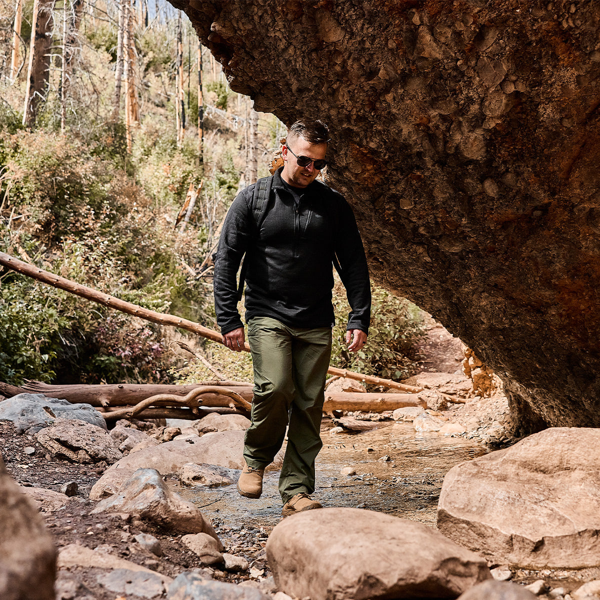 Wearing outdoor clothing and sunglasses, a person hikes along a rocky trail beneath a large rock formation. With their GORUCK Merino Challenge Socks - Crew providing friction resistance, they confidently traverse the forested area, surrounded by trees and bushes under a clear sky.