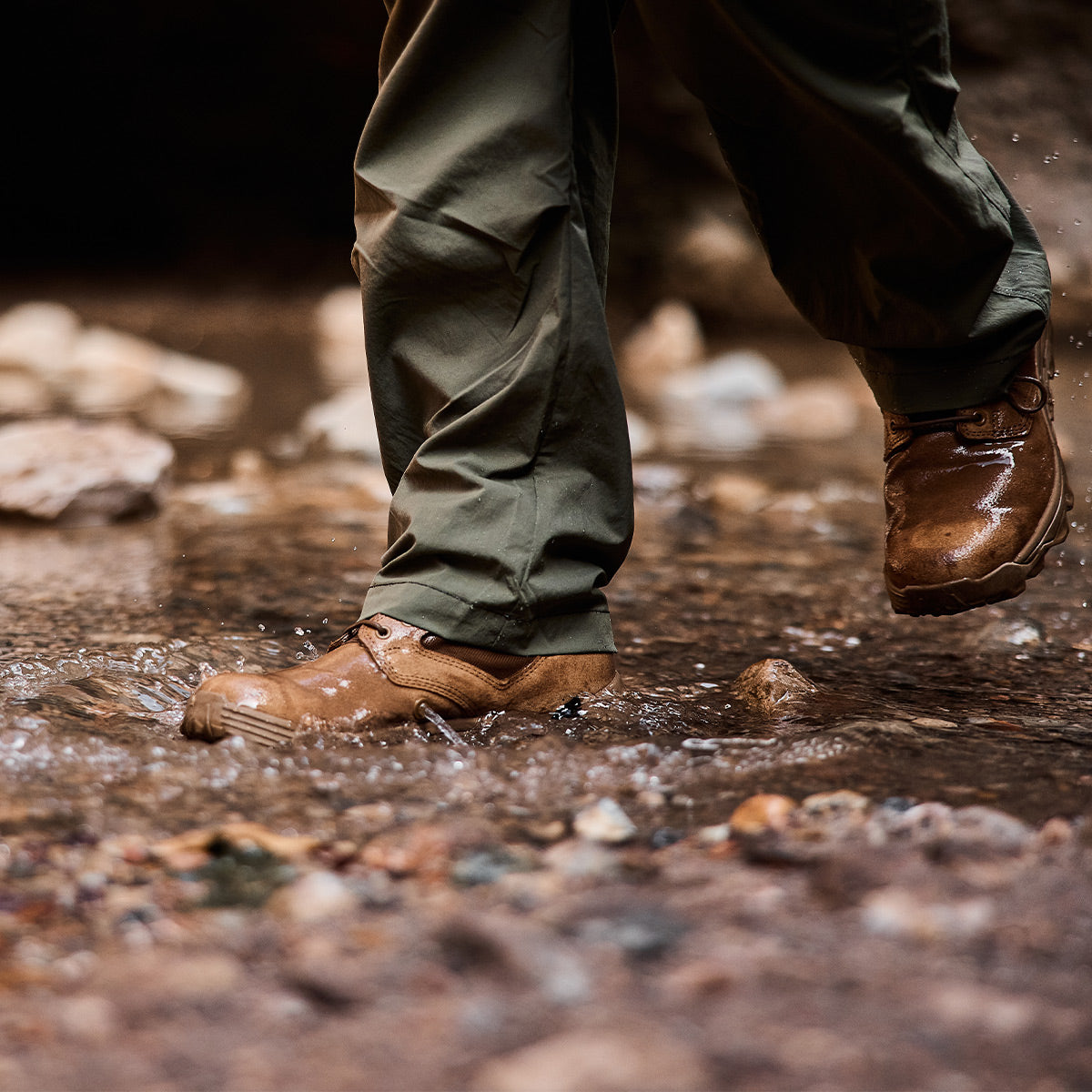 A person wearing Merino Challenge Socks - Crew and olive green pants strides through a shallow stream, splashing water and stepping on small stones, confident in their gear's durability thanks to the Scars Lifetime Guarantee from GORUCK.