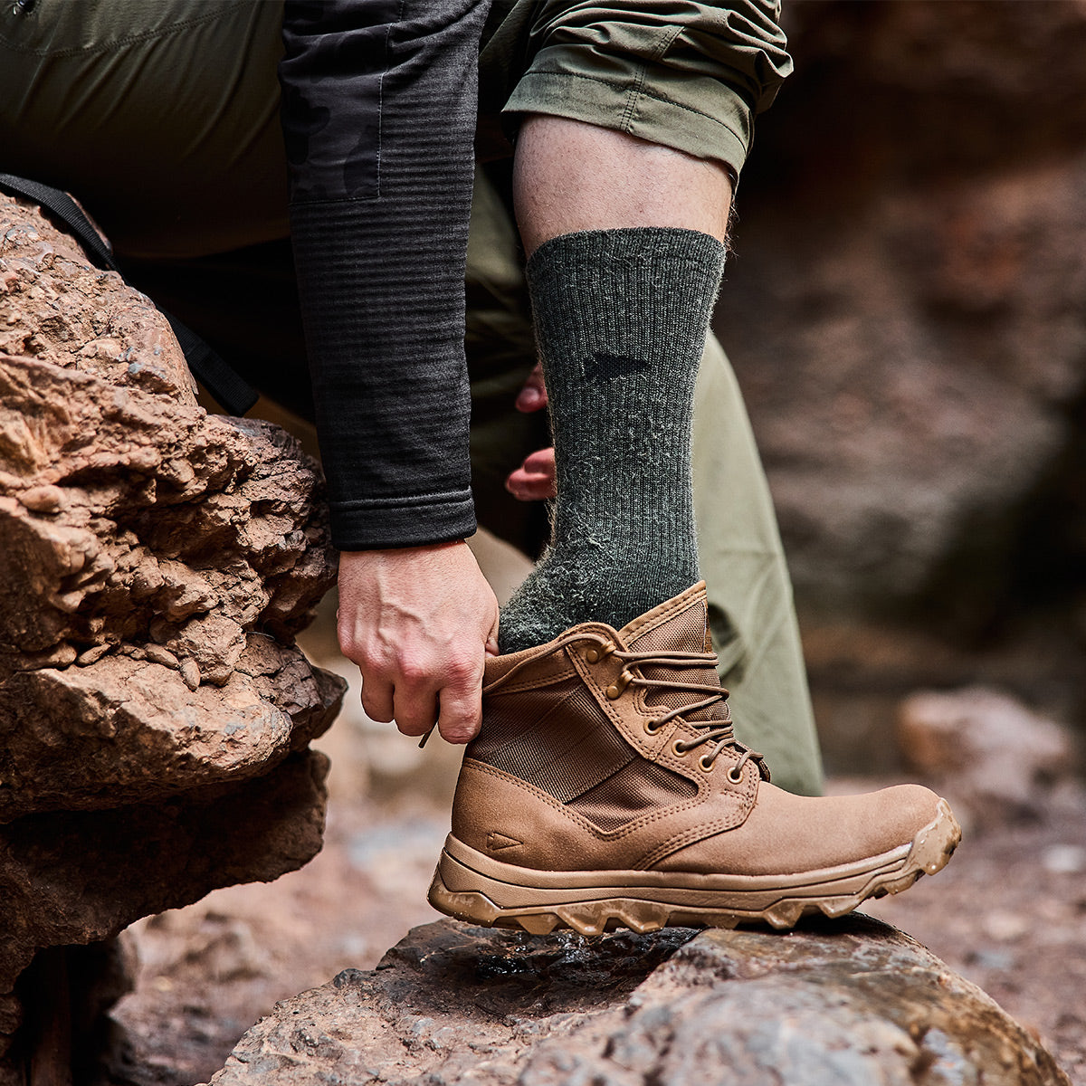 On a rocky trail, a person sitting on a stone adjusts their brown hiking boot, proudly Made in the USA. They are wearing GORUCK's Merino Challenge Socks - Crew in green and green cargo pants. A dark long-sleeve shirt is visible as well. The focus is on the boot and rugged terrain, equipped for any adventure with Scars Lifetime Guarantee.