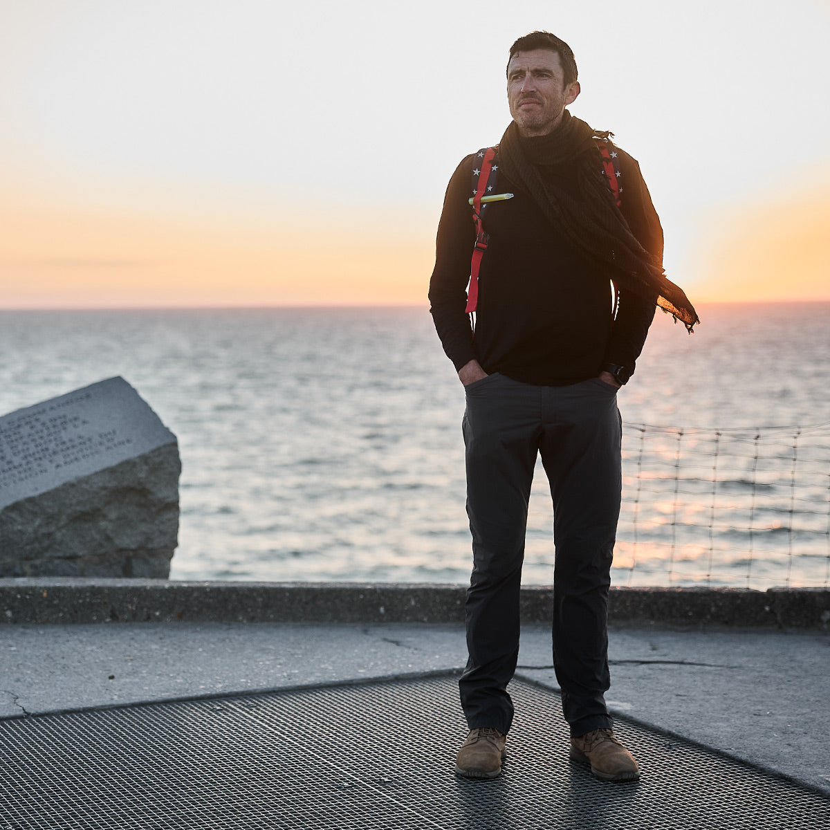 A man stands on a platform by the sea at sunset, wearing a backpack, scarf, and casual attire. He is sporting GORUCK's Merino Challenge Socks - Crew, known for their comfort and durability. To the left, a stone monument with inscriptions is visible, while a netted railing runs along the water's edge under a warm-toned sky.