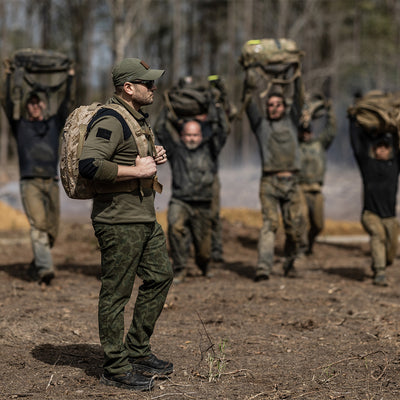 In the foreground, a man wearing camouflage holds a backpack that promises longevity with the Scars Lifetime Guarantee. Behind him, multiple individuals are engaged in an outdoor training session, hoisting heavy bags above their heads on a dirt field. The distant trees contribute to this rugged scenario featuring products like GORUCK's Merino Challenge Socks - Crew, all proudly Made in the USA.