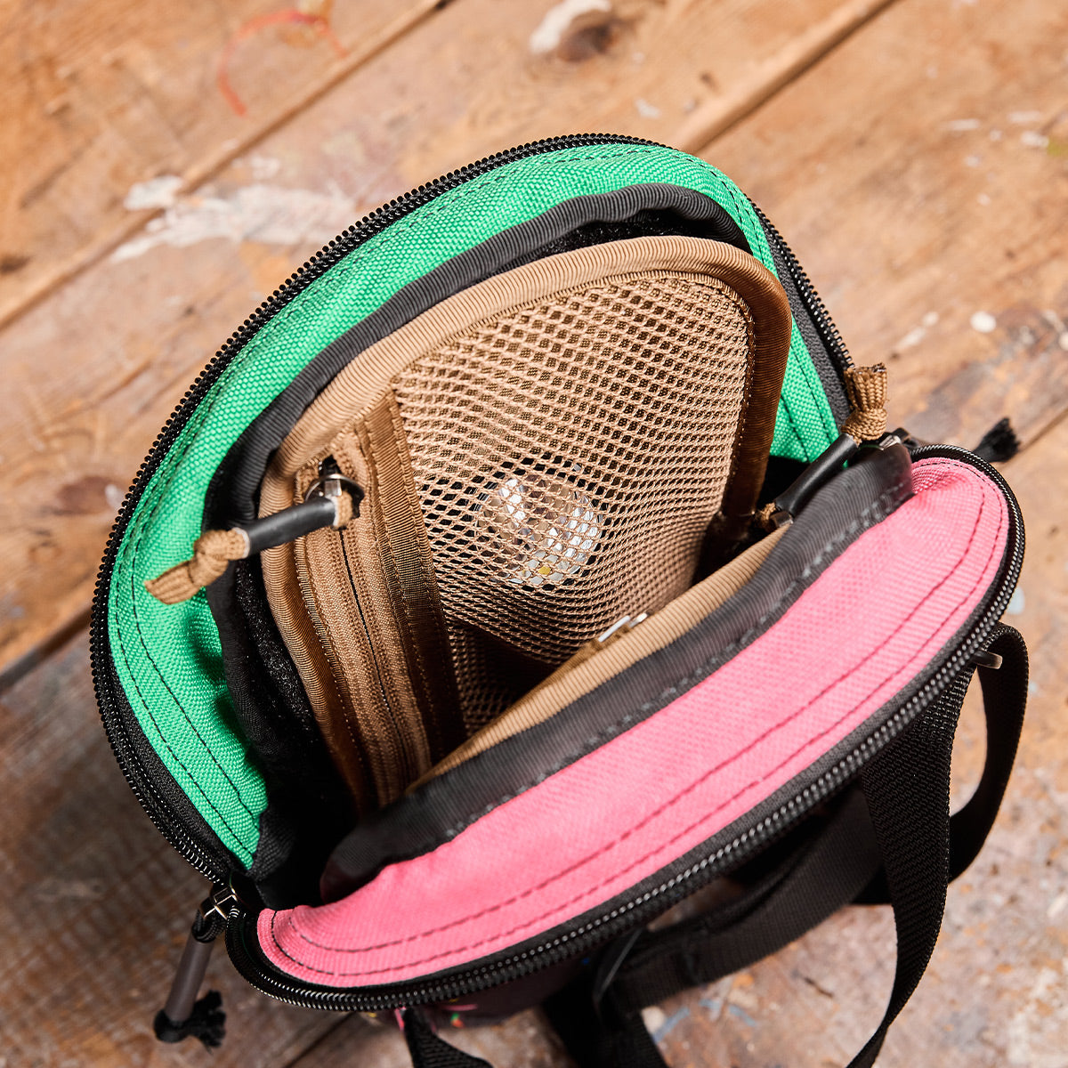 Open a multicolored GORUCK travel backpack on a wooden surface to reveal an Admin Panels brown mesh pocket inside, containing a small transparent plastic bag with a round object. The bag's green and pink sections are visible with black and brown detailing, perfect for EDC essentials.