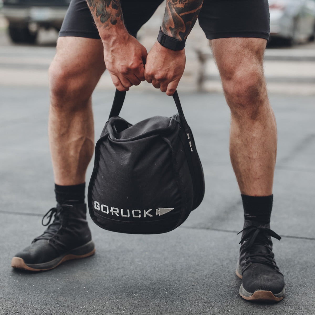 A person sporting GORUCK Men's Ballistic Trainers - Mid Top in Black + Gum with a Black Reflective Spearhead and black shorts grasps a GORUCK sandball bag crafted from CORDURA Ballistic Nylon. Their tattooed, muscular arms are prominent as they strike a pose on an outdoor gym floor.