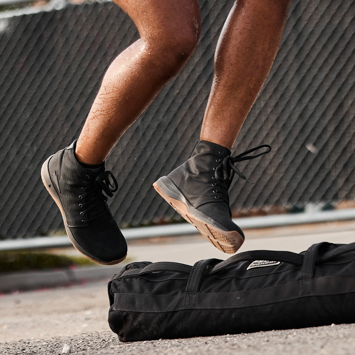 A close-up shot captures someone leaping on an outdoor surface, sporting the GORUCK Men's Ballistic Trainers - Mid Top in Black + Gum with a Black Reflective Spearhead, which are tailored for functional fitness. Nearby on the ground rests a black sports bag. In the background, a chain-link fence subtly showcases the sturdiness of CORDURA Ballistic Nylon in action.