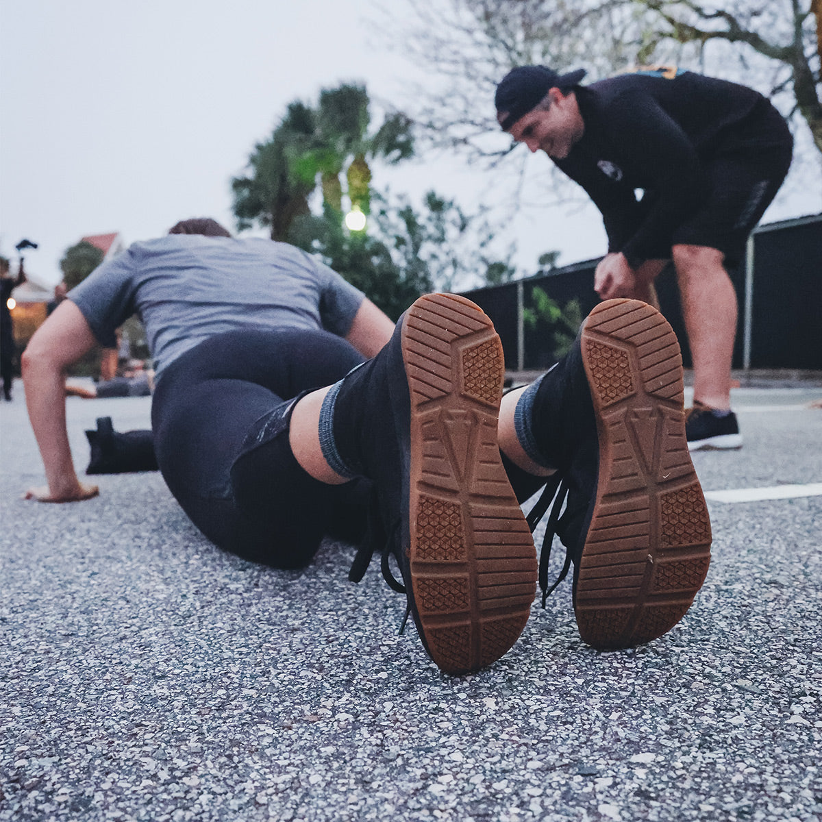 A person in athletic wear and Men's Ballistic Trainers - Mid Top - Black + Gum w/ Black Reflective Spearhead by GORUCK is doing push-ups on a paved surface, while another in a black outfit watches and encourages them. Trees and a fence are visible in the background, enhancing the scene's dynamic focus on strength and endurance.