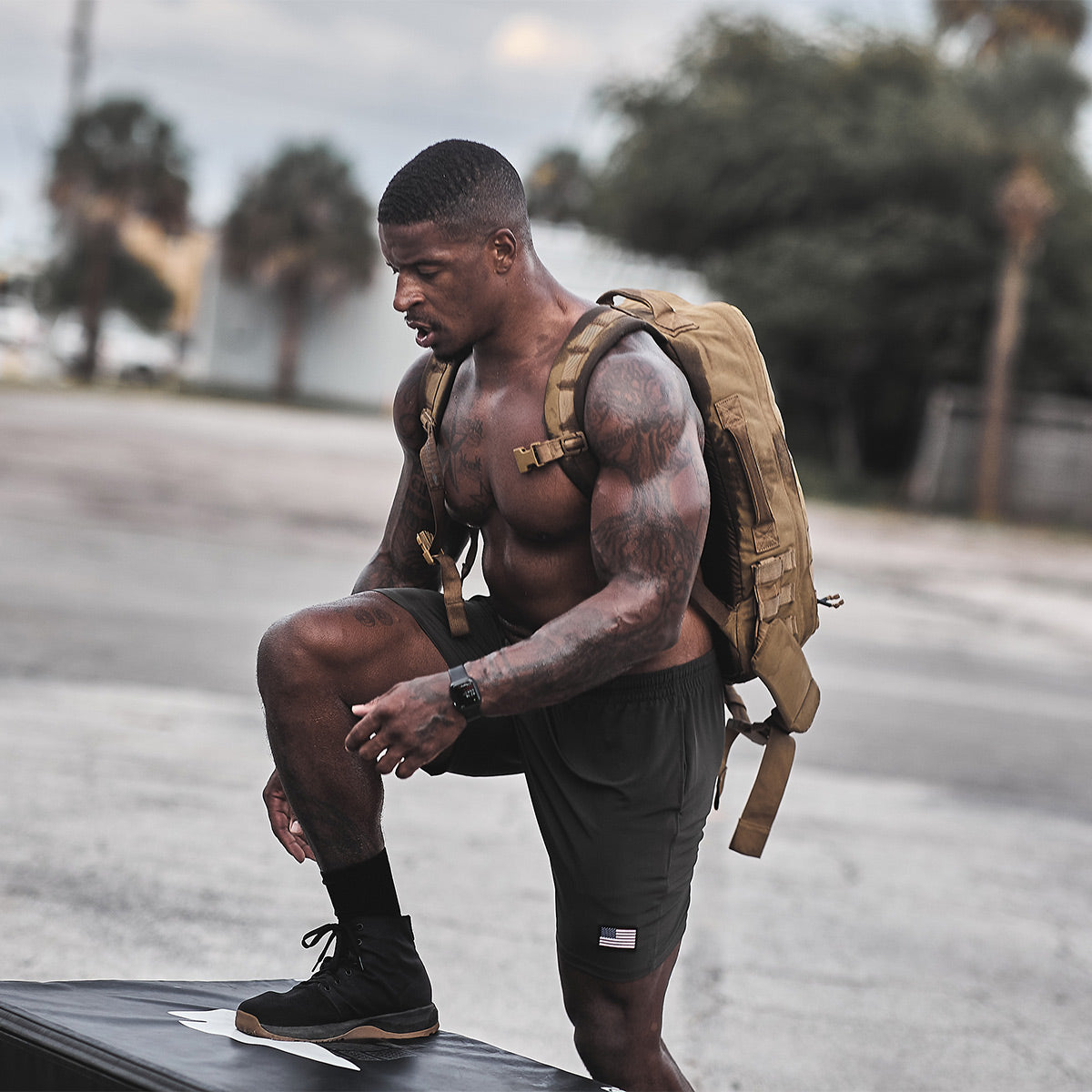 A muscular man with a backpack kneels on an outdoor platform, showcasing his GORUCK Men's Ballistic Trainers - Mid Top in Black + Gum with Black Reflective Spearhead. Shirtless in black shorts and sneakers, he highlights both style and the durability of CORDURA Ballistic Nylon against a backdrop of trees and buildings.