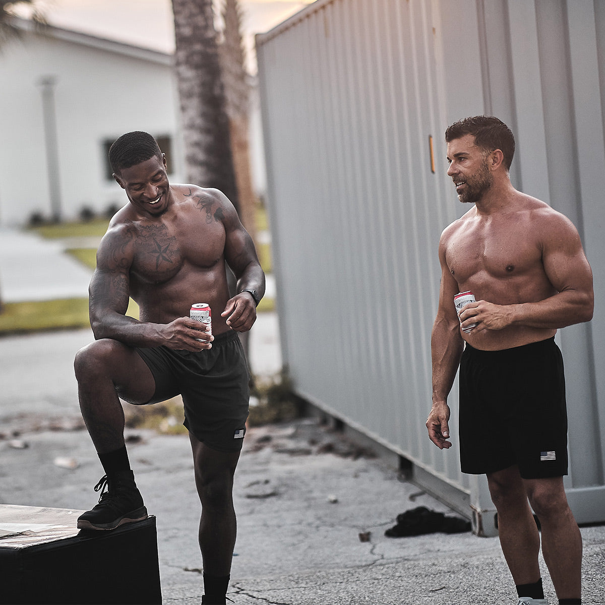 Two men with athletic builds are outdoors, each holding a drink. One man proudly shows off his CORDURA Ballistic Nylon shorts and smiles with his foot on a box labeled "3X Support." The other stands nearby. Both are shirtless and wearing GORUCK Men's Ballistic Trainers - Mid Top in Black + Gum with Black Reflective Spearhead, as a large container and palm trees set the scene.