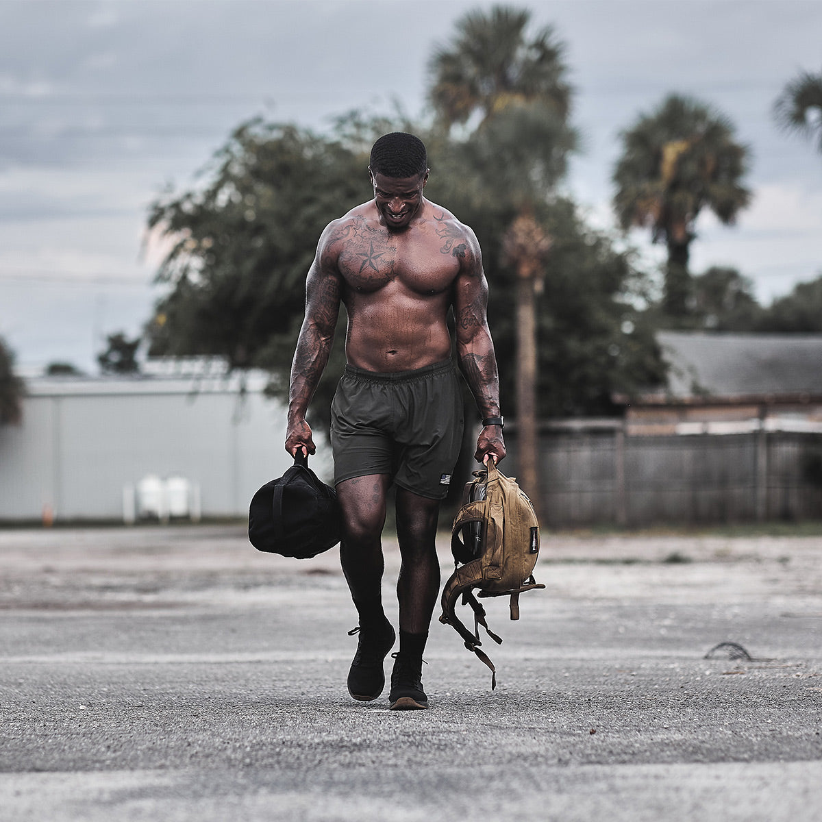 A muscular man walks down a street carrying a duffel bag and a backpack made of CORDURA Ballistic Nylon. He's shirtless, wearing gray shorts and the GORUCK Men's Ballistic Trainers - Mid Top - Black + Gum with Black Reflective Spearhead. The background features trees and buildings under an overcast sky.