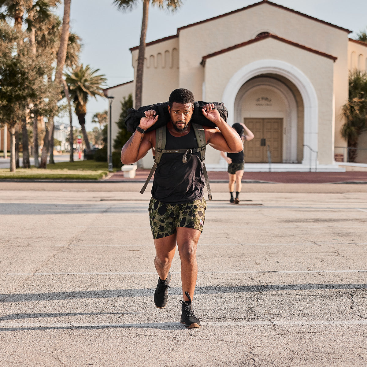 A man works out by carrying a sandbag on his shoulders as he walks on a paved surface. Sporting GORUCK Men's Ballistic Trainers - Mid Top in Black + Gum with a Black Reflective Spearhead, he is dressed in a black shirt and camouflage shorts. In the background, there is a building with an arched entrance and another person can be seen slightly behind him.