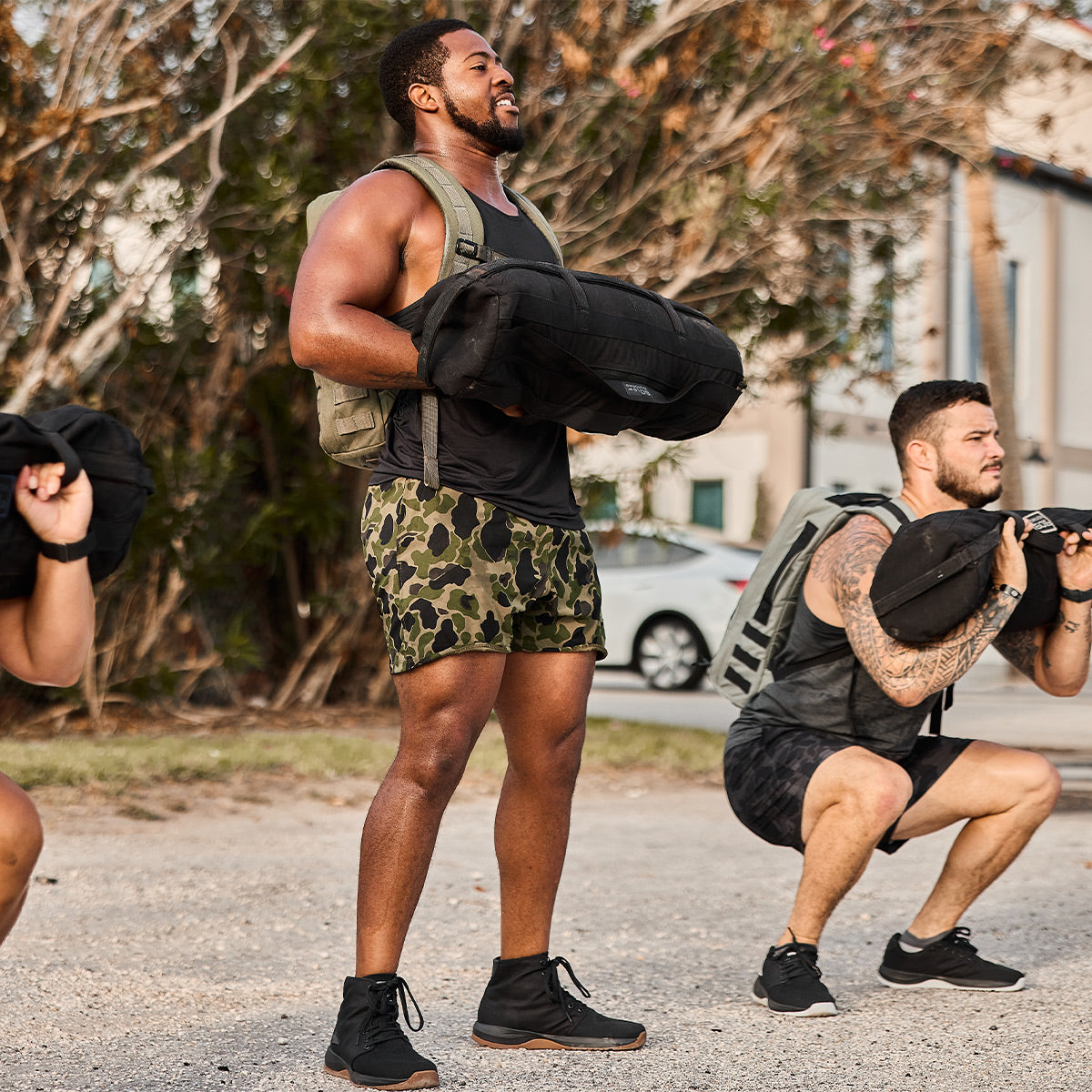 Three men are exercising outdoors in GORUCK's Men's Ballistic Trainers - Mid Top - Black + Gum with Black Reflective Spearhead, dressed in athletic gear. Two of them are squatting while wearing weighted backpacks crafted from CORDURA Ballistic Nylon, and the third is standing while holding a sandbag. They’re on a gravel surface surrounded by trees and buildings in the background.