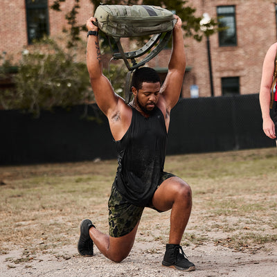 A man in a black sleeveless shirt and camouflage shorts, showcasing his GORUCK Men's Ballistic Trainers - Mid Top - Black + Gum with Black Reflective Spearhead, performs an overhead lunge with a camouflage bag outside. He kneels on one knee while holding the bag above his head, with a brick building and trees in the background.